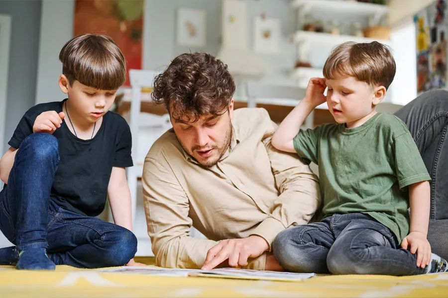 Dad and two young boys sat on a wooden floor, looking at a book together
