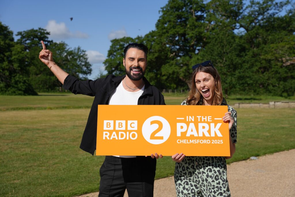 Rylan Clark and Ellie Taylor holding a BBC Radio 2 in the Park sign at Hylands Park